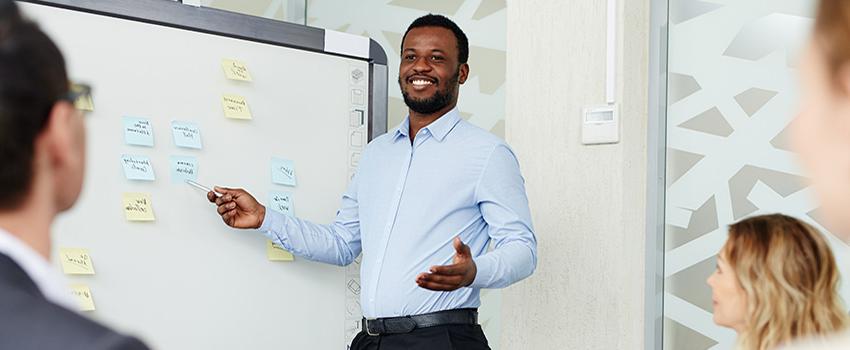 Man speaking in front of white board with people looking on.